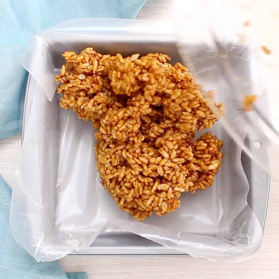 glass bowl tilted over lined baking pan dropping rice crispy treats mixture into pan