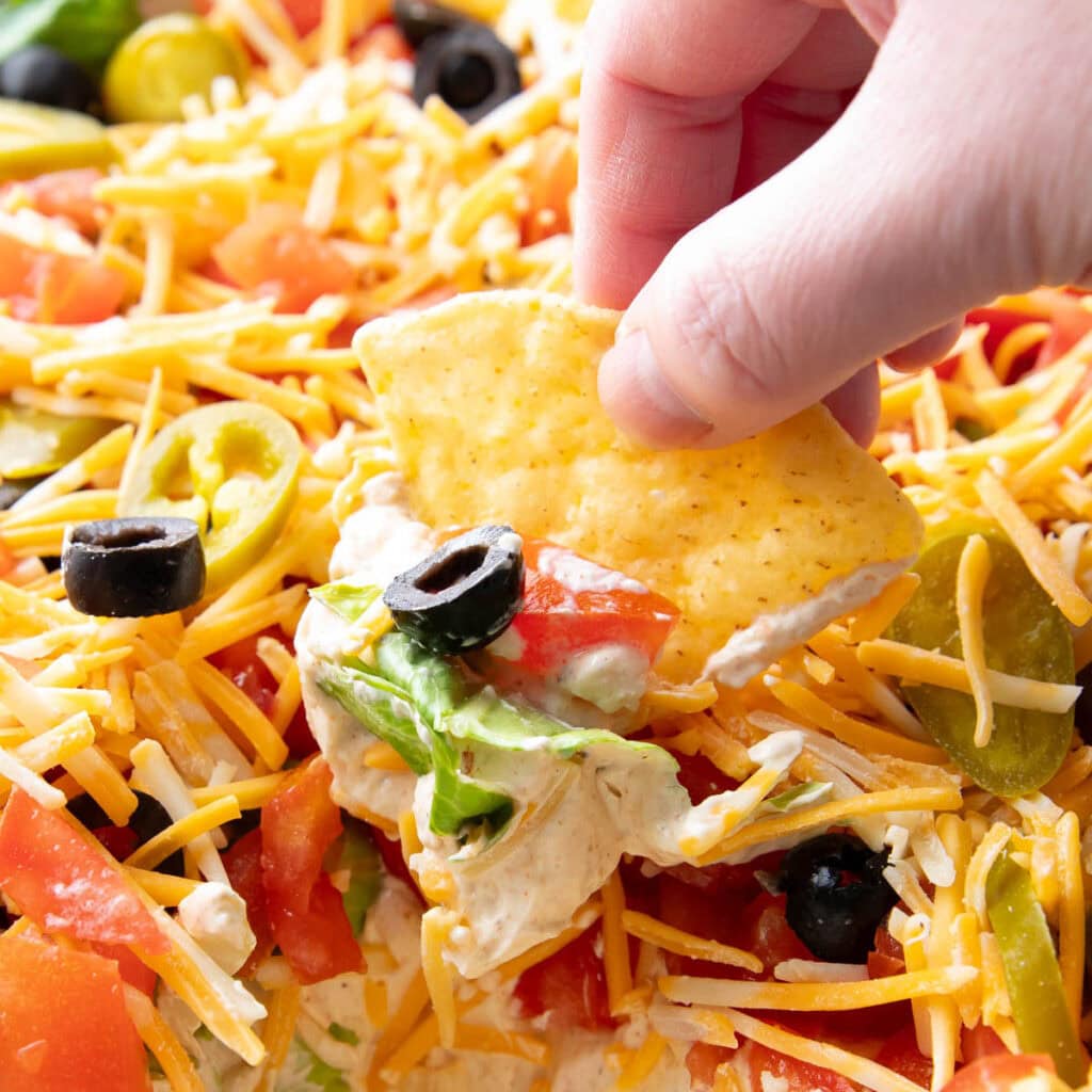 A hand dipping a tortilla chip into this layered taco dip and retrieving cream cheese seasoned dip with a black olive, lettuce, tomatoes, and cheese