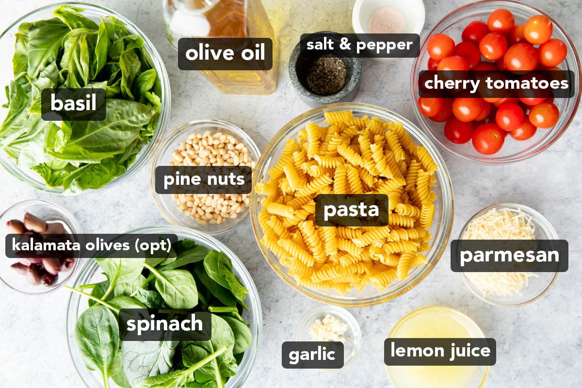 Tomatoes, pasta, spinach, basil, garlic and more ingredients for pesto pasta salad laid out on a table
