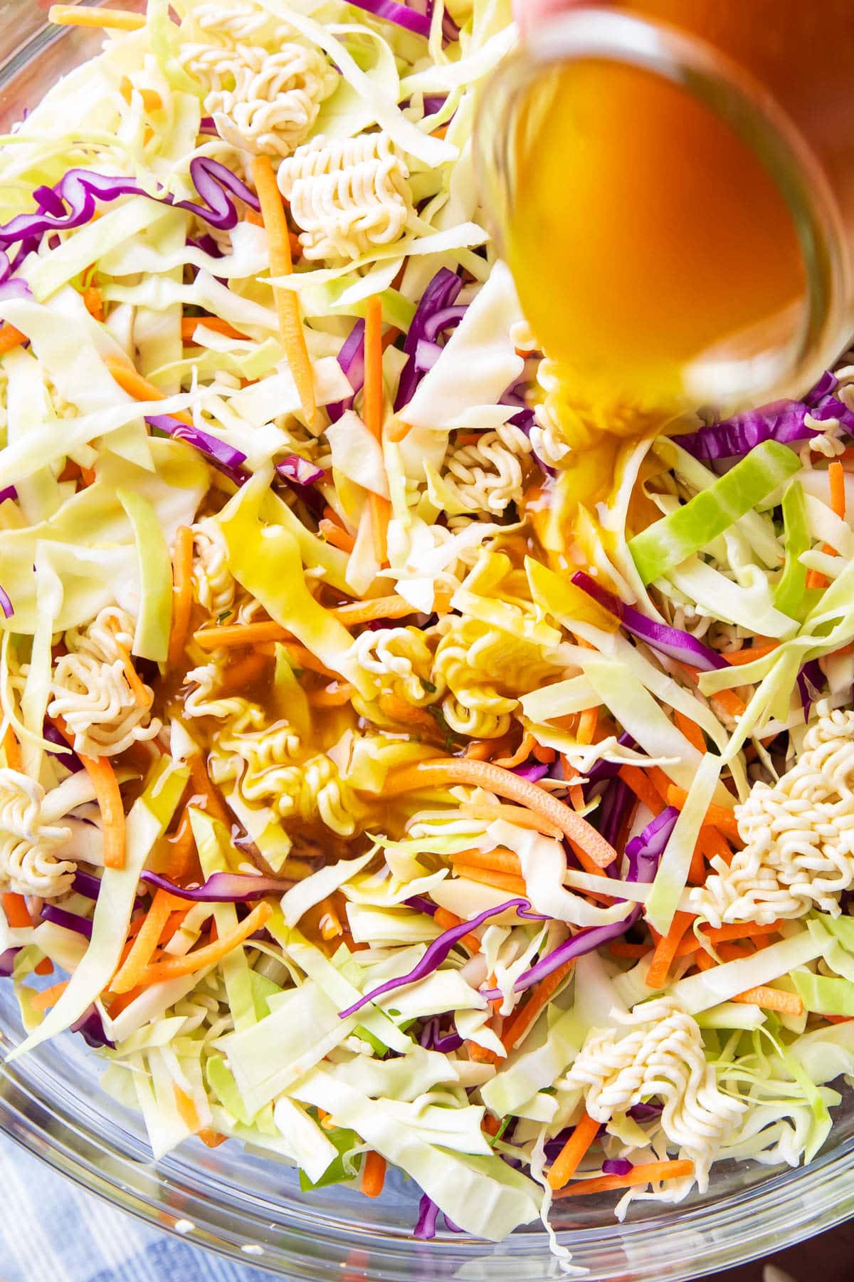Pouring a jar of homemade dressing over ramen noodle salad packed with cabbage, green onions, almonds, and carrots.