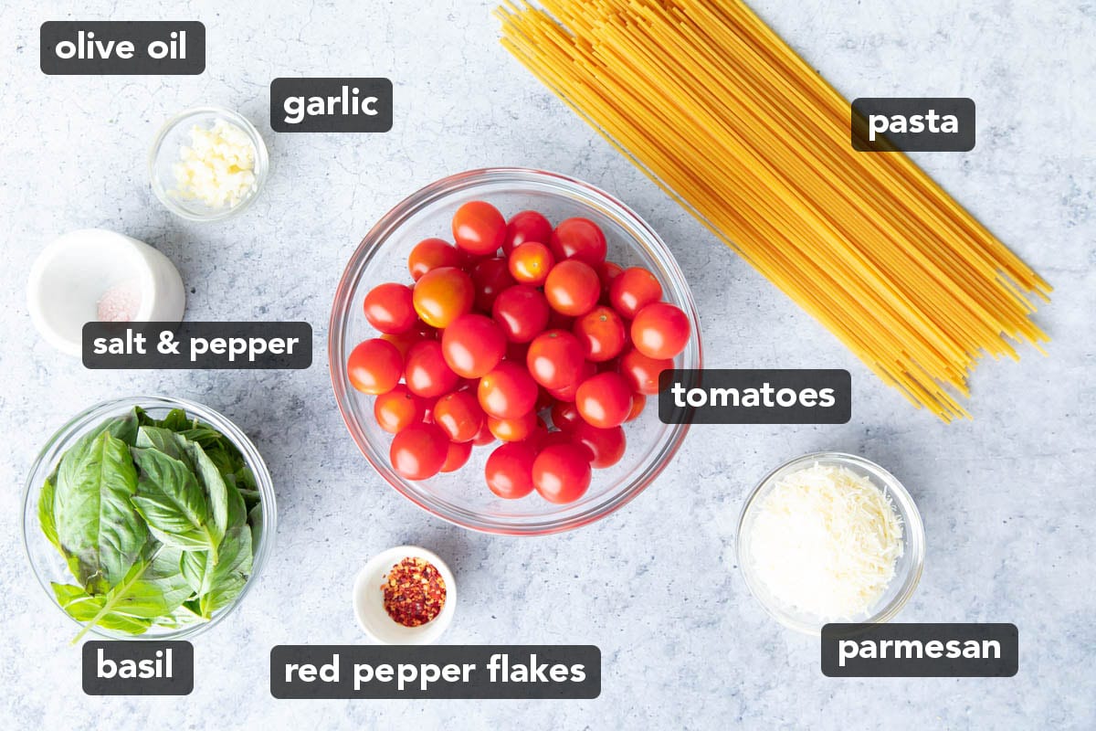 Tomato Basil Pasta ingredients including spaghetti, cherry tomatoes, red pepper flakes, fresh basil, parmesan and more laid out on a table